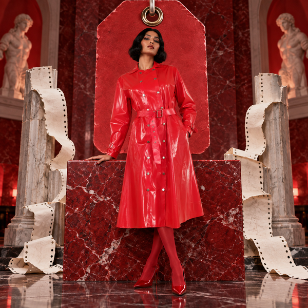 Adult performer in glossy red belted trench coat before a monumental scarlet coat-check ticket and marble columns. ARTPOP CHAOS v2 Avant-Garde Art Editorial.