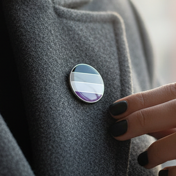 Asexual Pride Badge on a dark gray coat lapel, featuring horizontal stripes in gray, white, and purple. A hand with dark nails touches the lapel, symbolizing empowerment and prideful self-expression.