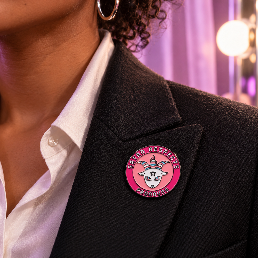 Pink Satan Respects Pronouns enamel pin attached to a black blazer lapel.