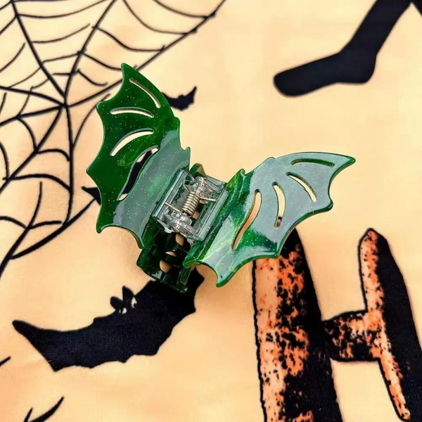 The Gothic Glam Bat Hair Claw, a green glittery hair accessory shaped like bat wings, rests on Halloween-themed fabric, embodying bold, queer self-expression and empowering style with its elegant and edgy design.