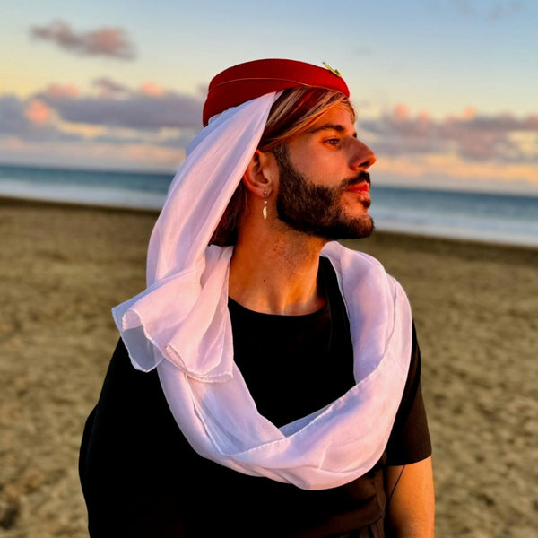 A bearded person on a beach at sunset wears the First Class Purser Statement Headwear with a red cap and white draped cloth, exuding bold queer elegance and high-fashion self-expression.