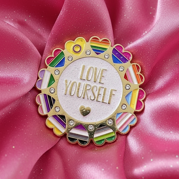 The Love Yourself Rainbow Enamel Pin shines on pink satin, featuring a gold border with rhinestones, rainbow heart accents, and LOVE YOURSELF in gold on white—a proud declaration of LGBTQIA+ pride and self-love.