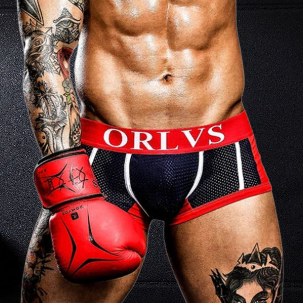 A fit, tattooed man showcases ORLVS Mesh Pocket Boxers, revealing muscular legs and abs. Red-and-black mesh with a bold waistband embodies queer pride and sexy self-expression.