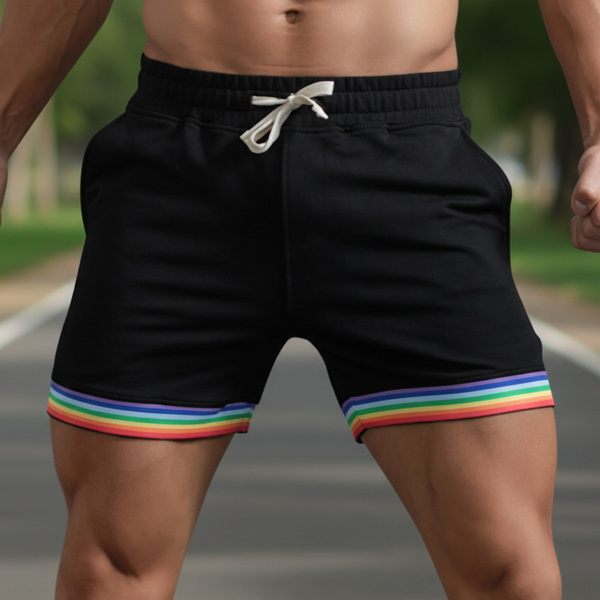 A queer person stands on a path in black Rainbow Sprint Sports Shorts with rainbow-striped trim and white drawstring; trees blur behind.