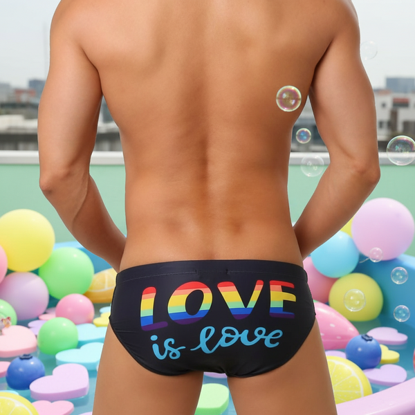 Person's bare back wearing Love Is Love swim briefs with rainbow lettering, surrounded by pastel balloons and foam hearts. Celebratory, empowering swimwear showcasing queer pride and self-expression in a joyful setting.
