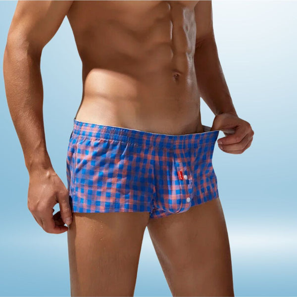 A shirtless man showcases the SEOBEAN Bold Wear Distressed Boxer Briefs, featuring a blue-and-red checkered pattern and distressed design. He pulls the waistband to display the fit, exuding queer confidence and self-expression.