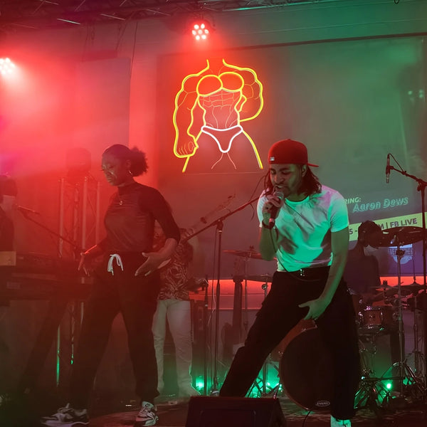 Yellow neon outline of a muscular person in briefs glowing behind performers onstage