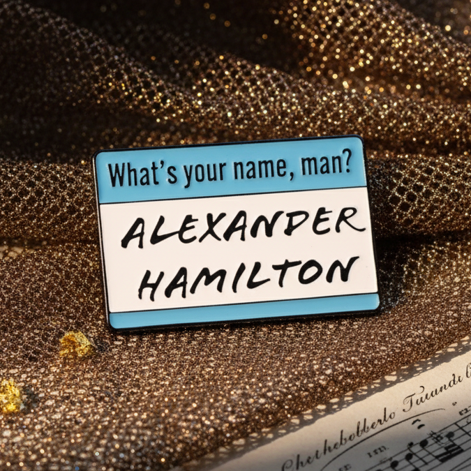 What’s My Name? Alexander Hamilton Enamel Pin on sparkly gold fabric, showcasing sky-blue and white name-tag design. Celebrates queer pride and self-expression with bold confidence.