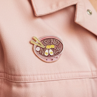 Send Noods Enamel Pin on pink jacket—depicts a vibrant ramen bowl with noodles and chopsticks. Celebrate LGBTQIA+ pride and self-expression with this bold, empowering accessory from Queer In The World.