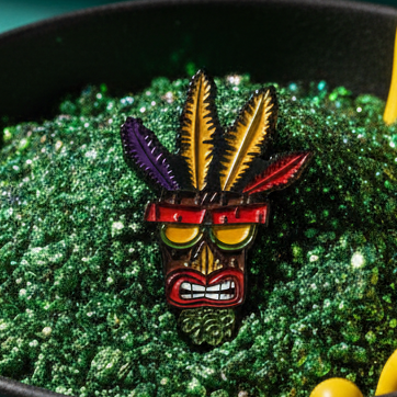 Close-up of Aku Aku Crash Bandicoot enamel pin with colorful feathers and red sunglasses