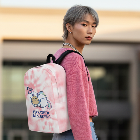Minimalist backpack with “I’d Rather Be Sleeping” slogan and a snoozing cat, worn by a short silver-haired person in a pink sweater outdoors; blurred building in background.