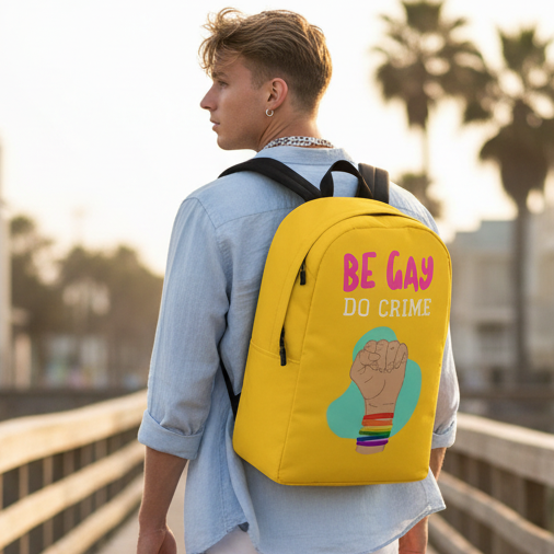 Be Gay Do Crime bright yellow backpack with slogan above a fist in rainbow bracelets, worn by a young man on a sunny boardwalk.
