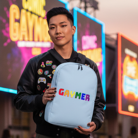 Gaymer Minimalist Backpack, light blue with "GAYMER" in rainbow lettering. Queer gamer in patched black jacket among lively crowd, bright signs.