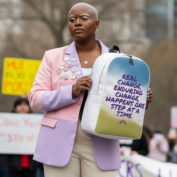Person holding a white-and-olive backpack reading “REAL CHANGE, ENDURING CHANGE HAPPENS ONE STEP AT A TIME”
