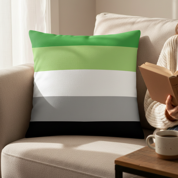 Aromantic pride pillow with green, white, gray, and black stripes on a beige sofa; person in cream sweater reads nearby.