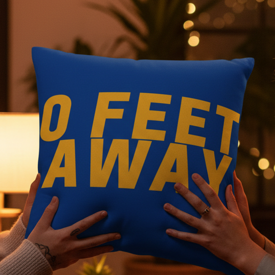 Two hands hold the Zero Feet Away Grindr Premium Pillow with bold yellow “0 FEET AWAY” text, warm light and blurred fairy lights.