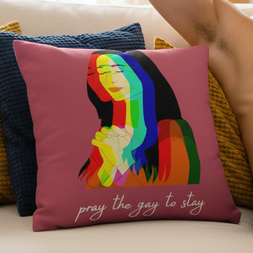 Pink "Pray The Gay To Stay" pillow with rainbow illustration of a praying person, white cursive text, on blue pillow and beige sofa.