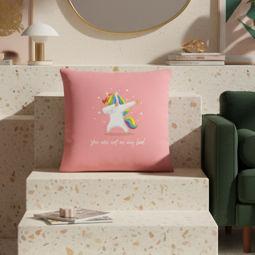 Pink "You Are Not On My Level" LGBT pillow with rainbow-maned unicorn on terrazzo steps; green sofa, white book, mirror, lamp nearby.