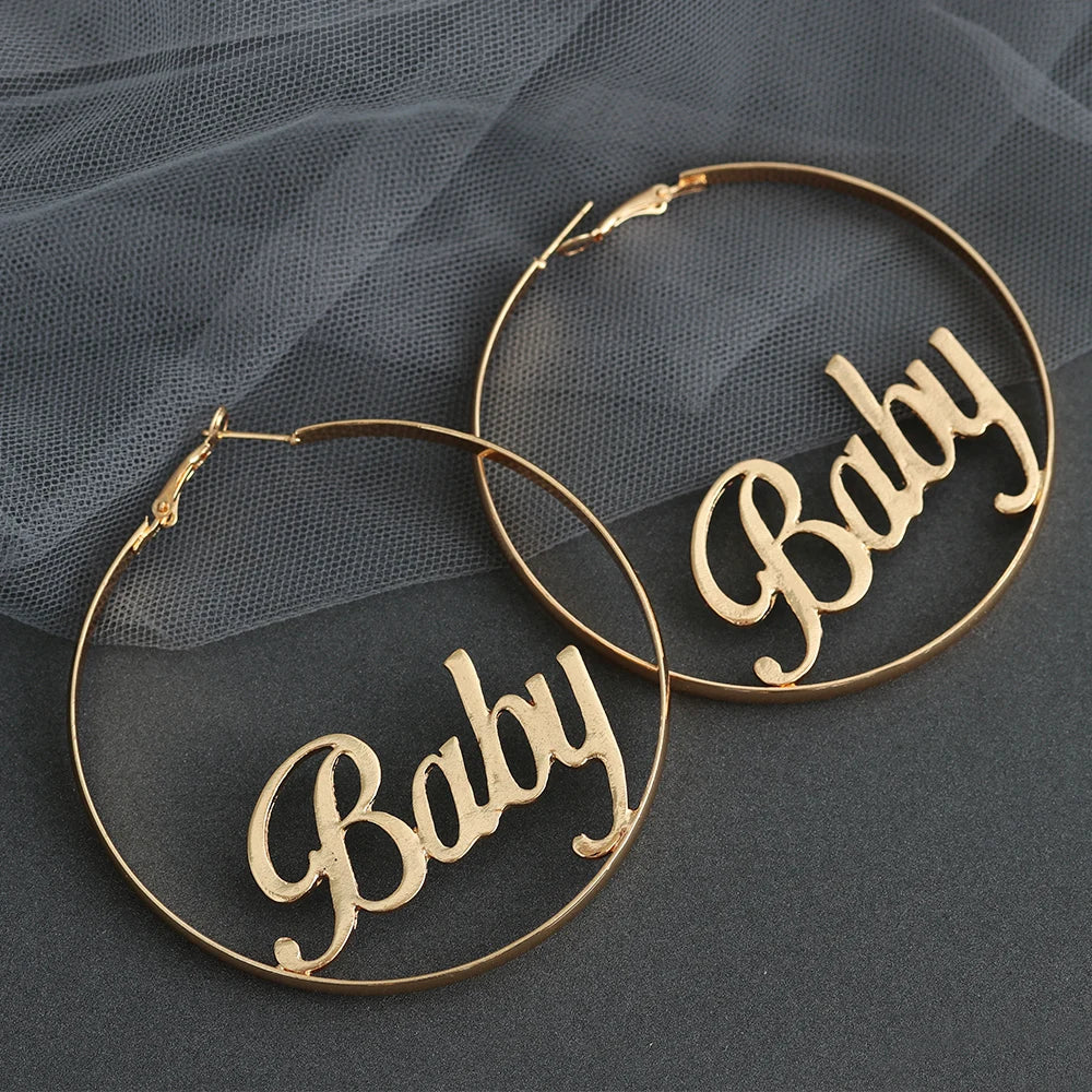 Gold BABY hoop earrings with cursive lettering, laid flat on dark gray fabric
