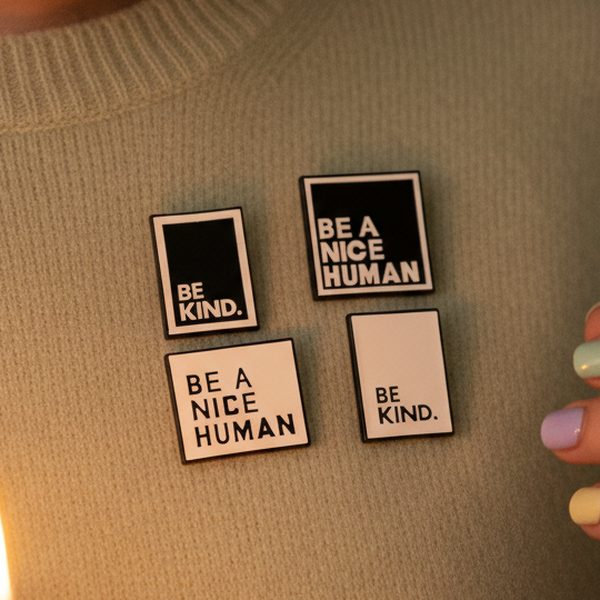 Four square enamel pins reading “BE A NICE HUMAN” and “BE KIND.” on fabric