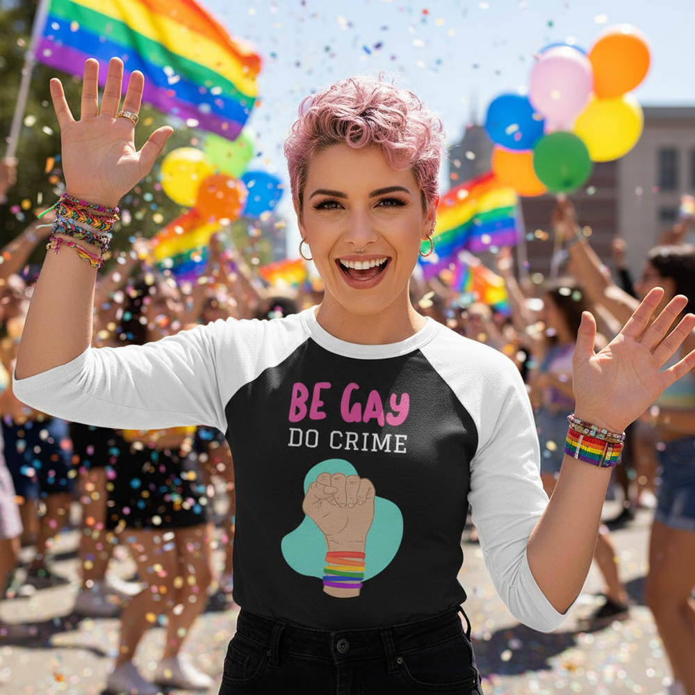 “Be Gay Do Crime” 3/4 sleeve raglan shirt worn by a smiling person with pink hair and rainbow bracelets