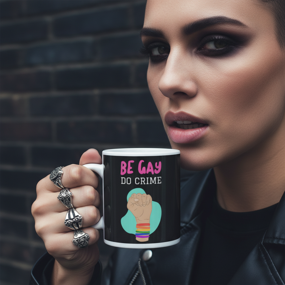 Person holds a black mug reading “BE GAY DO CRIME” with a raised fist graphic