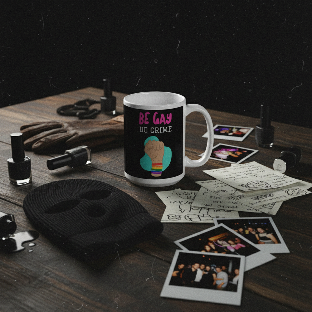 White mug reading “BE GAY DO CRIME” with a raised fist and rainbow stripes on a prop-filled table