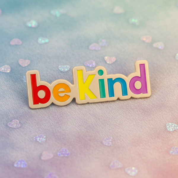 “be kind” rainbow enamel pin on a pastel heart-patterned background