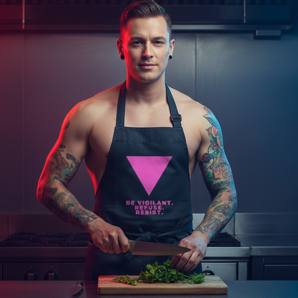 Tattooed person wearing a black apron reading “BE VIGILANT. REFUSE. RESIST.” while chopping greens