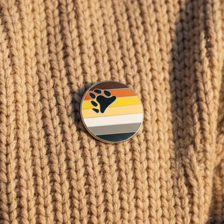 Bear pride enamel pin with black paw print and brown, tan, yellow, cream, gray, and black stripes