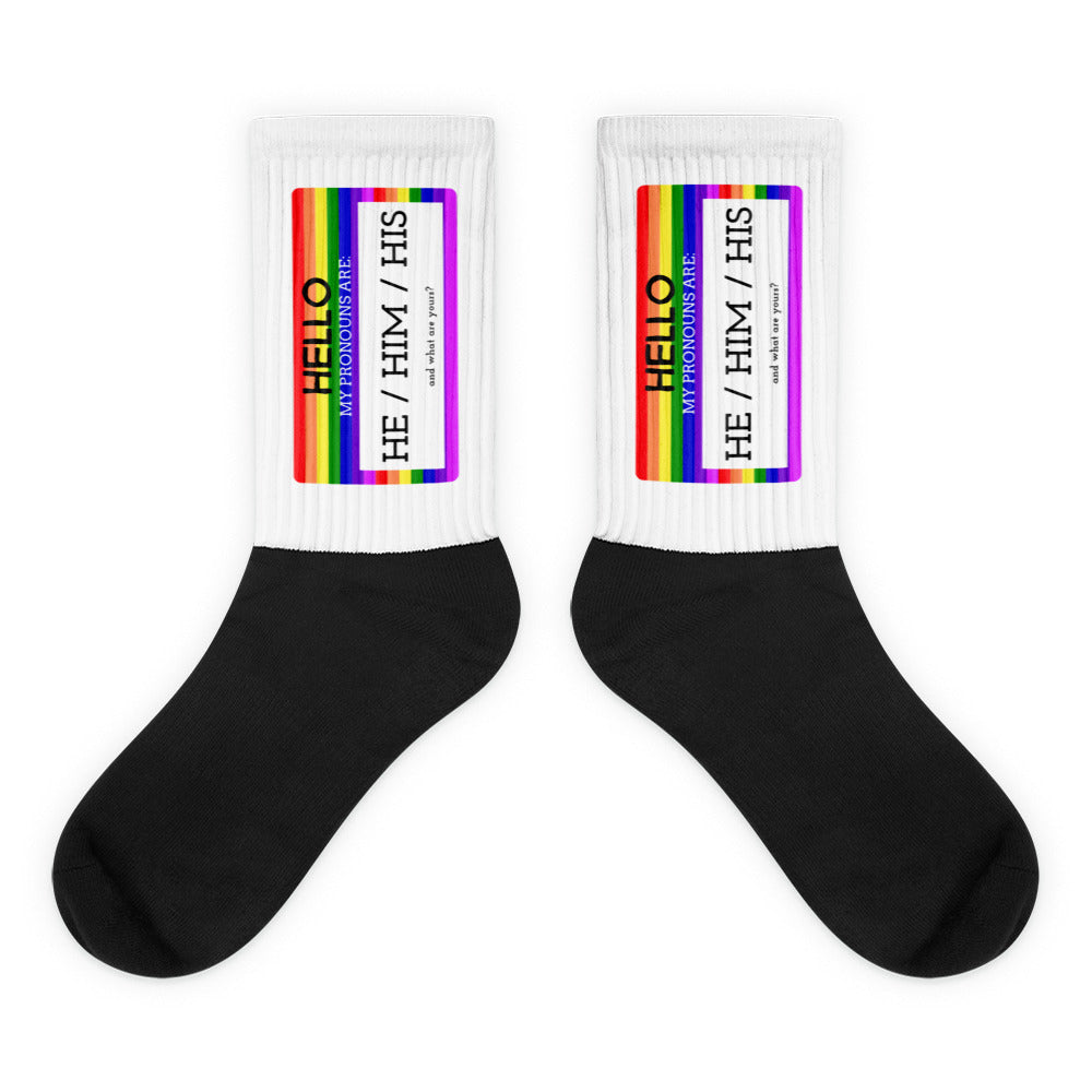 Pair of black socks with white cuffs and rainbow labels reading “HE / HIM / HIS.”