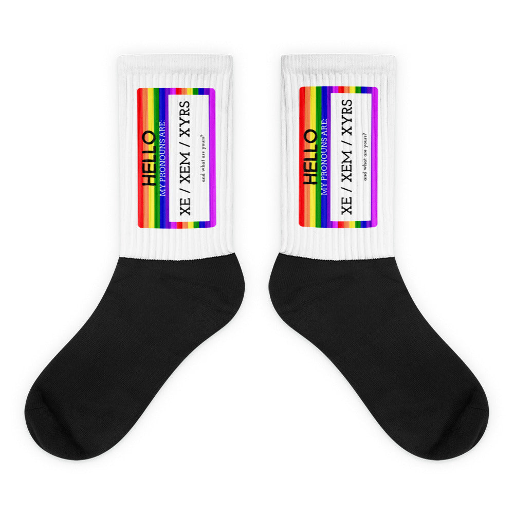 Pair of black-and-white socks with rainbow cuffs labeled “Hello, my pronouns are Xe / Xem / Xyrs.”