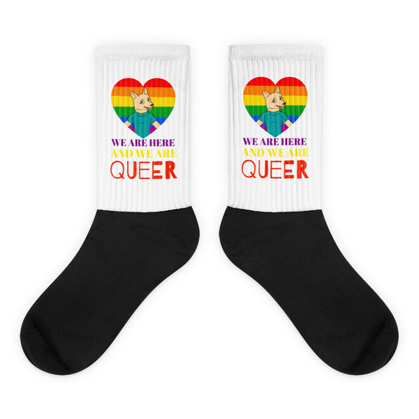 We Are Here And We Are Queer Socks feature a rainbow heart, corgi character, and pride message on white cuffs with black feet, celebrating LGBTQIA+ visibility and self-expression.