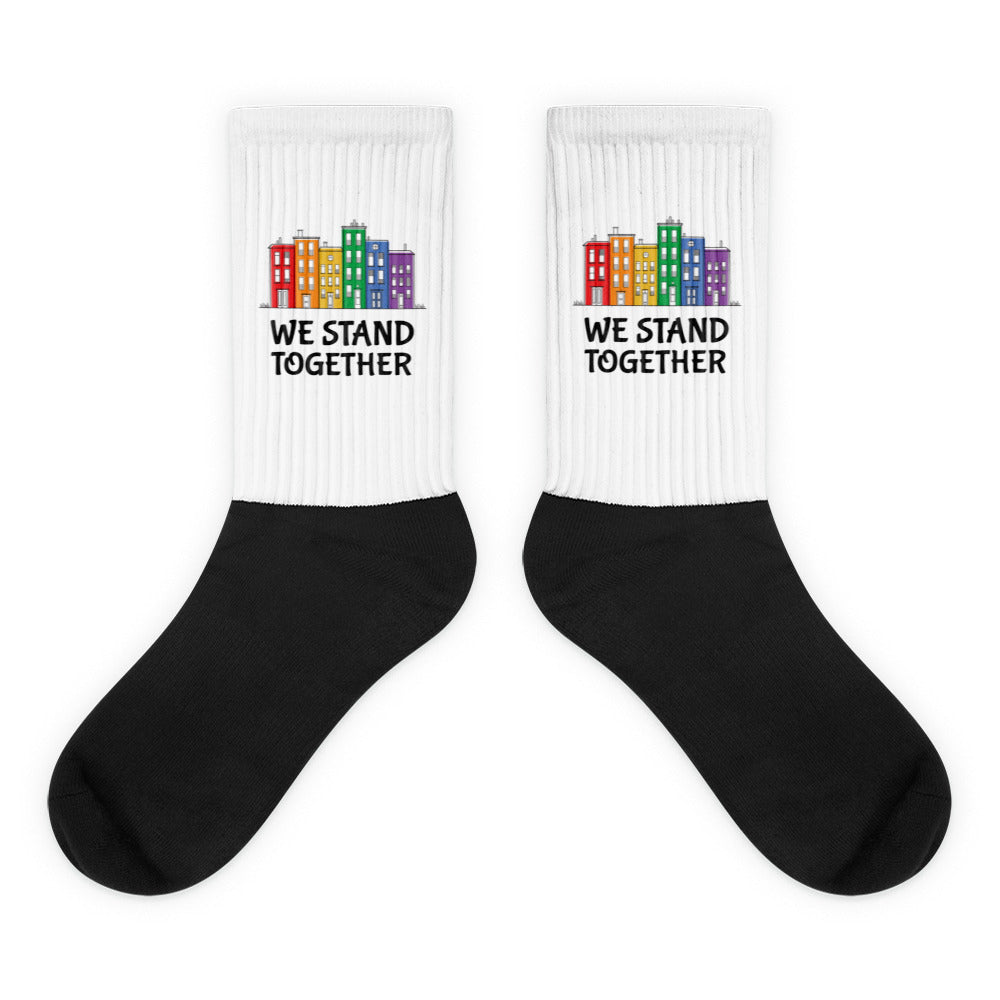 Pair of crew socks with black feet, white cuffs, rainbow buildings, and “WE STAND TOGETHER”