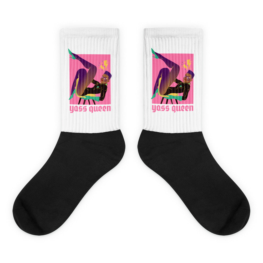 Pair of socks with black feet and white cuffs reading “yass queen” below colorful pop-art figures