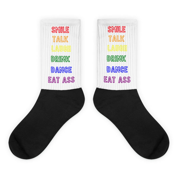 Smile, Talk, Laugh, Drink, Dance, Eat Ass Socks feature bold black feet and white cuffs with vibrant, rainbow-colored words, embodying joyful self-expression and queer pride in a comfortable, stylish crew-length design.
