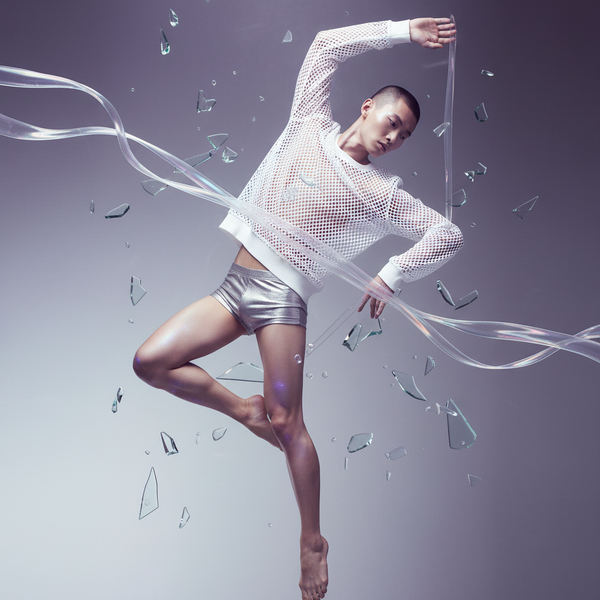 Model floating in a white mesh long-sleeve shirt and metallic shorts amid ribbons and glass shards