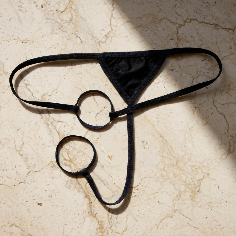 Black strappy men's underwear with two circular rings laid flat on ivory stone.