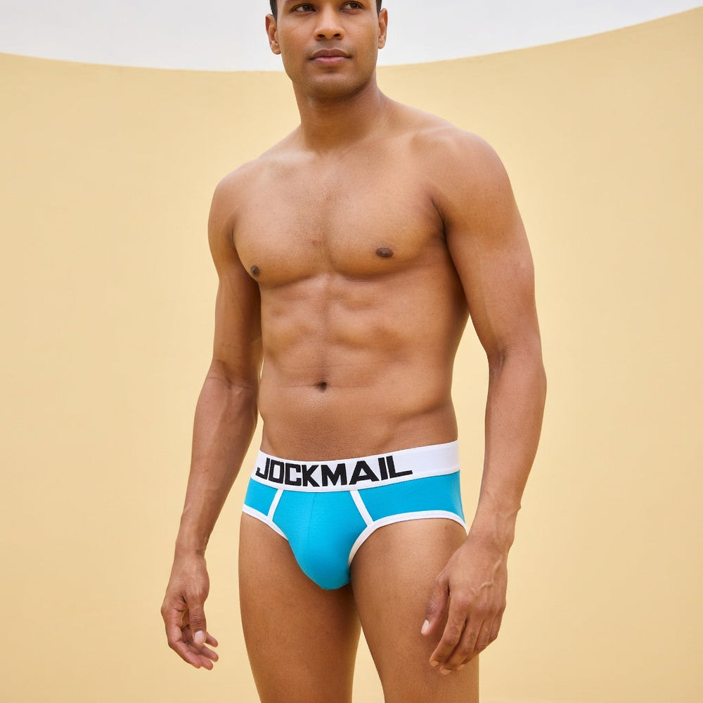 Adult model wearing blue Jockmail backless briefs with white piping against a yellow wall.