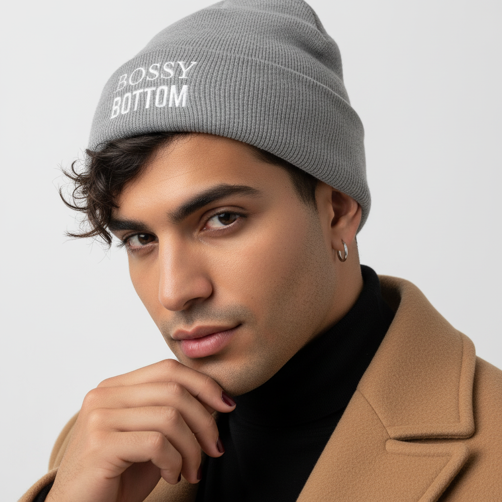Gray "Bossy Bottom" Cuffed Beanie on wavy-brown-haired person in tan coat, hoop earring, dark nails; white backdrop.