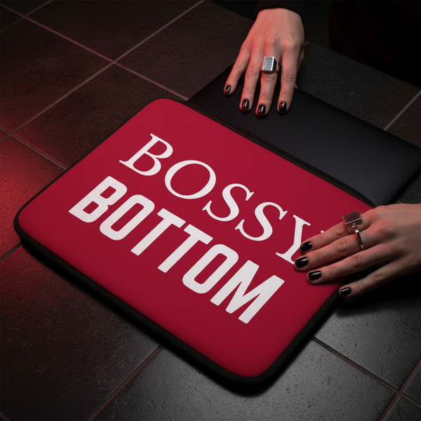 Bossy Bottom Laptop Sleeve with bold white text, shown under hands with black nail polish and silver rings on dark tiles.