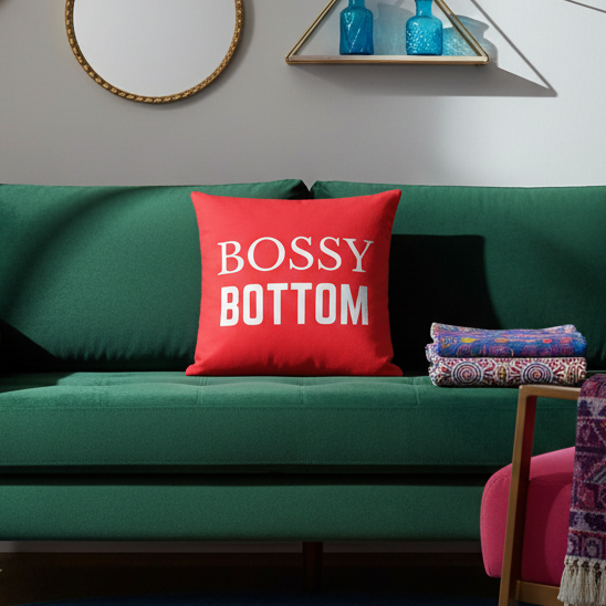 The “Bossy Bottom” pillow with bold white text on bright red sits on a green sofa, near patterned blankets, mirror