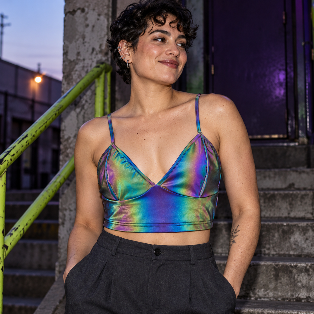 Queer model wearing an iridescent rainbow reflective crop top with thin straps on concrete steps outside a club.
