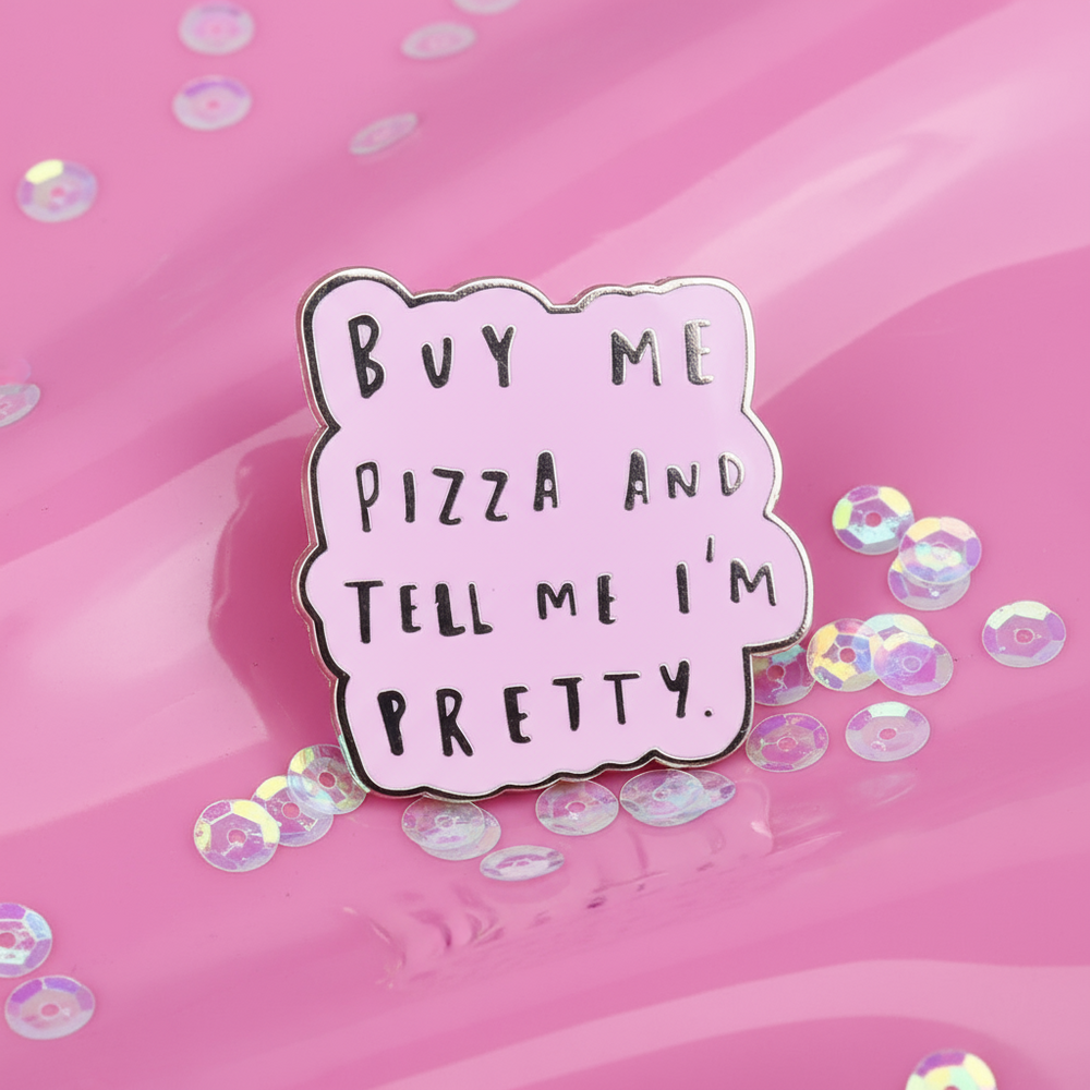 Pink enamel pin reading “BUY ME PIZZA AND TELL ME I'M PRETTY” on a pink surface