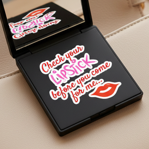 Black compact mirror on beige surface shows a vinyl “Check your lipstick before you come for me...” sticker in pink, red