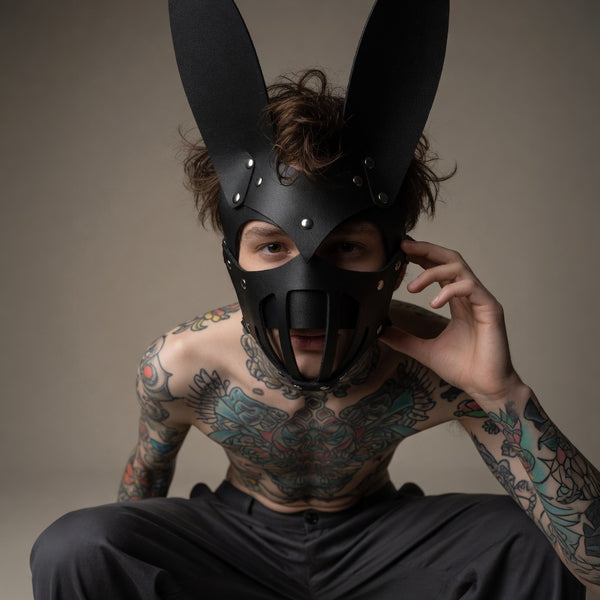 Tattooed model wearing a black bunny-eared BDSM mask with metal studs