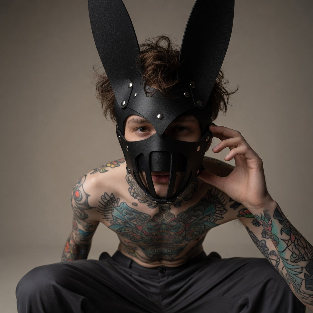 Tattooed model wearing a black bunny-eared BDSM mask with metal studs