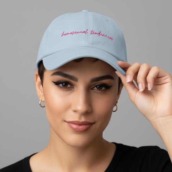 Light blue embroidered dad hat with “Homosexual Tendencies” in pink script, held by a queer person against a gray backdrop.