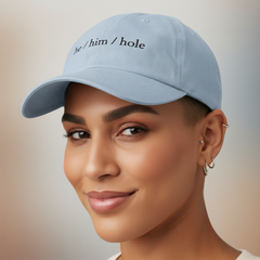 Light blue “He / Him / Hole” cap with black embroidery, worn by a person smiling gently, multiple silver hoops, soft blurred background.