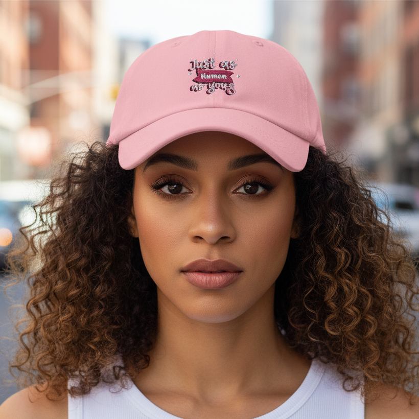 Pink cotton "Just As Human As You" queer cap with decorative text, worn by a young woman outdoors on a city street.